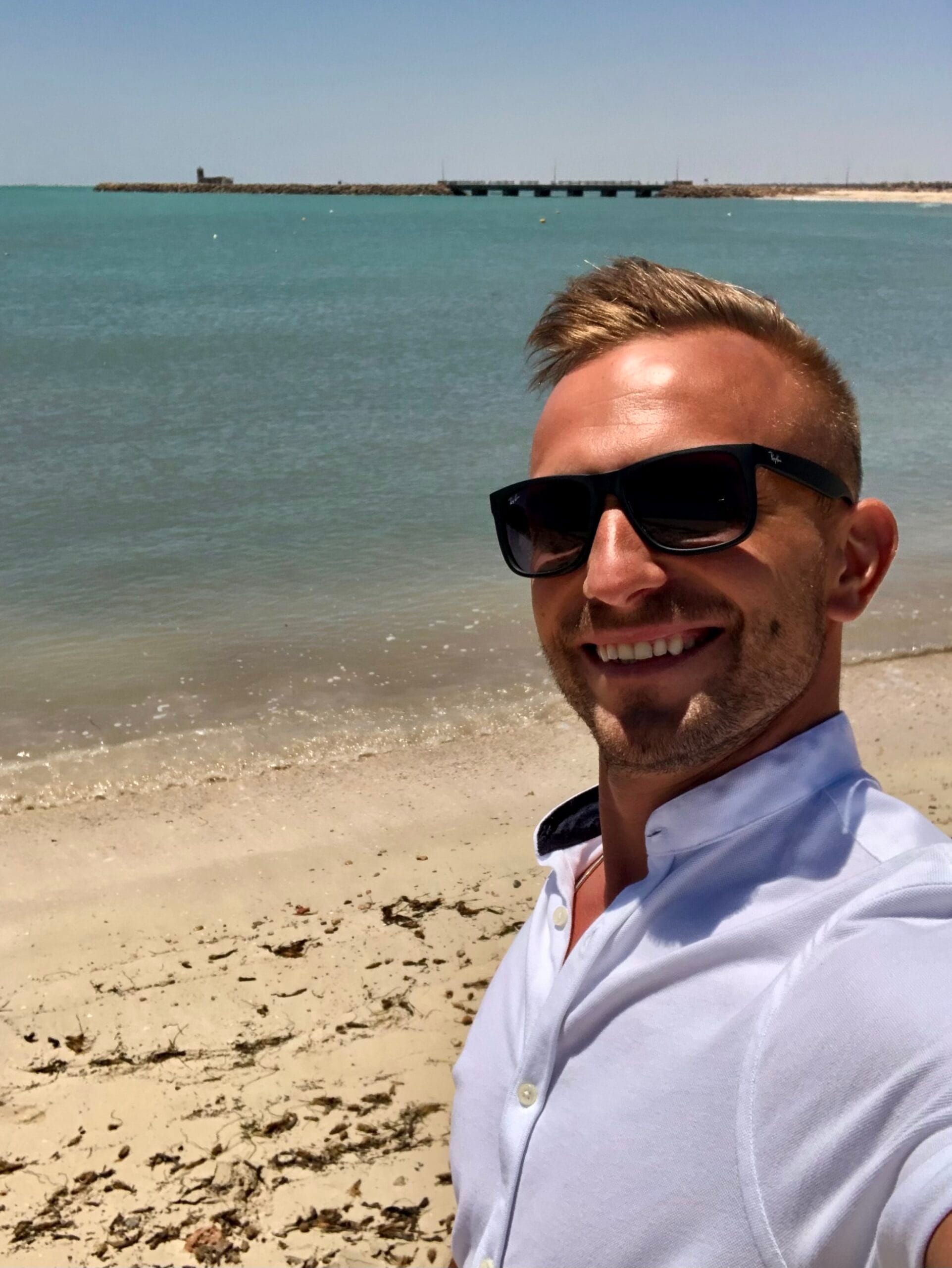 A man smiling in a white shirt on a sunny beach, used as a profile photo on the About Me page.